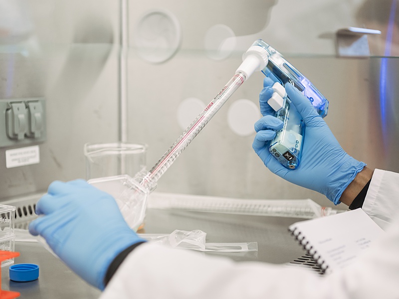 A person wearing blue gloves and a lab coat uses a pipette to transfer liquid into a container inside a laboratory setting, with lab equipment and a notebook visible in the background.
